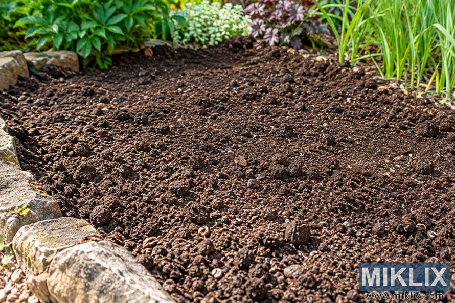 Freshly prepared dark garden soil in a stone-edged bed, surrounded by green and purple foliage and ready for bulb planting. Freshly prepared dark garden soil in a stone-edged bed, surrounded by green and purple foliage and ready for bulb planting.