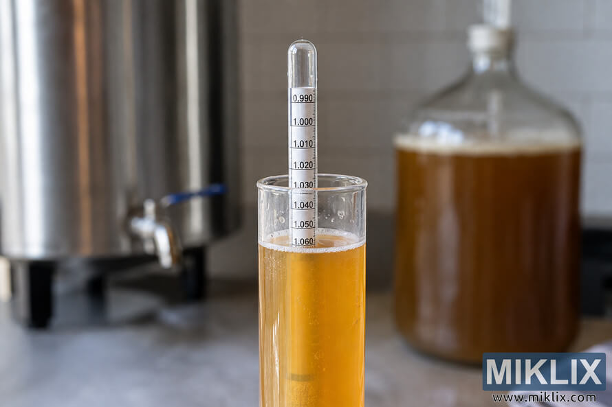 Close-up of a glass hydrometer in a golden beer sample, with a stainless-steel vessel and glass fermenter blurred in the background.