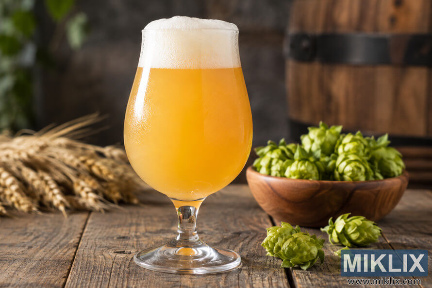 Hazy golden Belgian-style ale with a creamy white head in a tulip glass on a rustic wooden table, surrounded by fresh green hops and golden grain, with a blurred wooden barrel in the background. Hazy golden Belgian-style ale with a creamy white head in a tulip glass on a rustic wooden table, surrounded by fresh green hops and golden grain, with a blurred wooden barrel in the background.