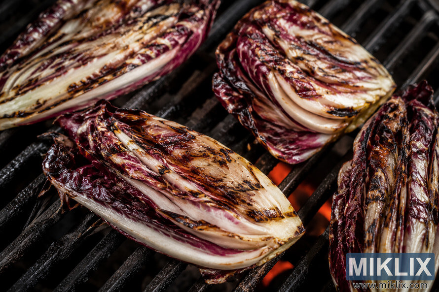Close-up of grilled radicchio wedges resting on a hot barbecue grill, showing deep caramelization, dark char marks, purple and white leaves, and glowing embers beneath the grates. Close-up of grilled radicchio wedges resting on a hot barbecue grill, showing deep caramelization, dark char marks, purple and white leaves, and glowing embers beneath the grates.