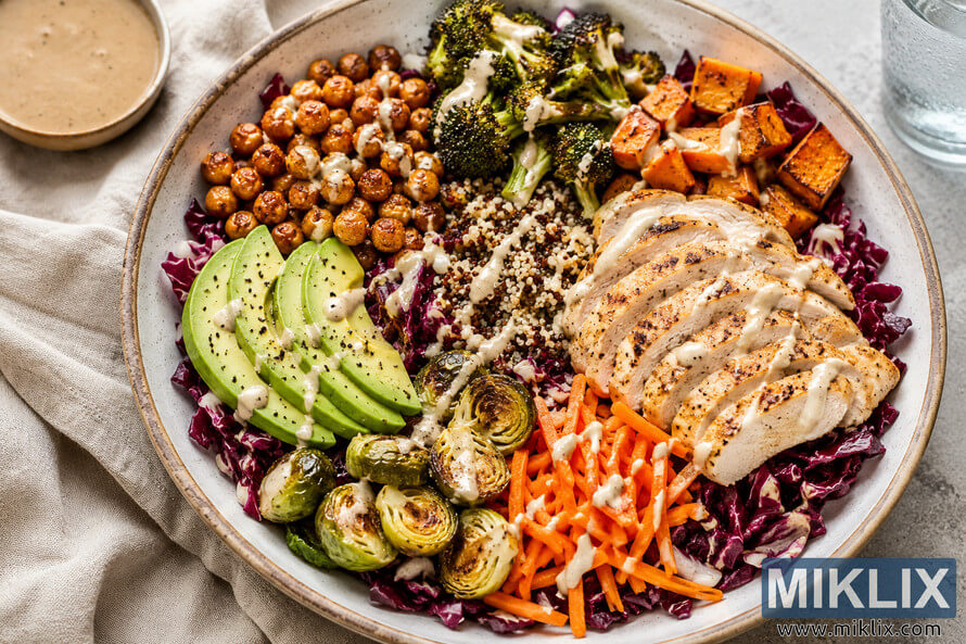 Landscape food photograph of a ceramic Buddha bowl built on chopped radicchio and topped with grilled chicken, quinoa, roasted chickpeas, broccoli, sweet potato, Brussels sprouts, avocado, shredded carrots, and tahini dressing. The bowl sits on a beige linen cloth over a gray stone surface with a small bowl of dressing and a glass of water nearby. Landscape food photograph of a ceramic Buddha bowl built on chopped radicchio and topped with grilled chicken, quinoa, roasted chickpeas, broccoli, sweet potato, Brussels sprouts, avocado, shredded carrots, and tahini dressing. The bowl sits on a beige linen cloth over a gray stone surface with a small bowl of dressing and a glass of water nearby.