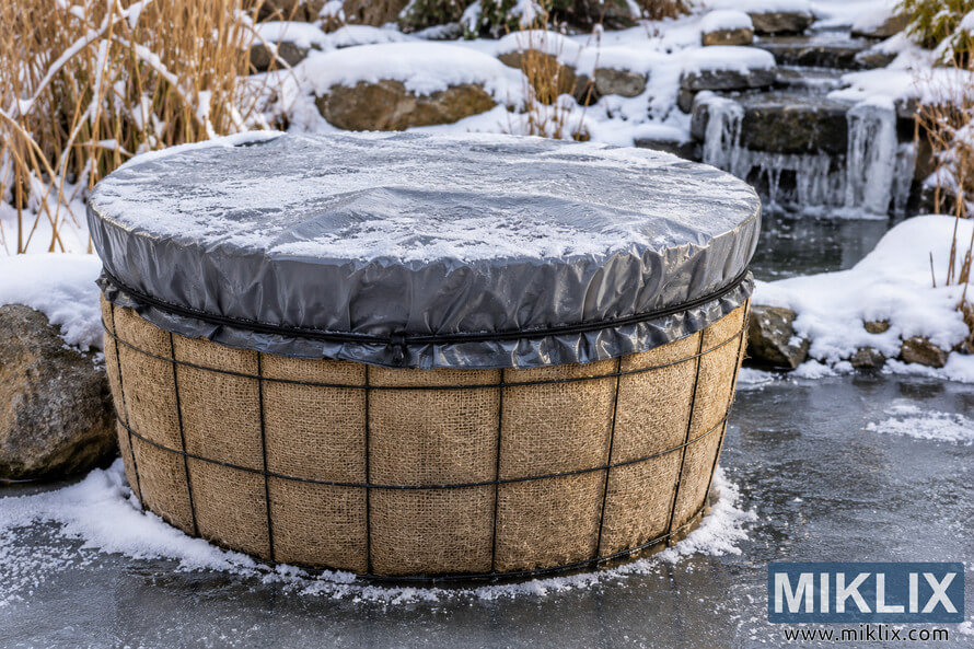 Large garden container wrapped in burlap and wire mesh with a frost-covered gray tarp, surrounded by snow, ice, rocks, and a frozen pond waterfall.
