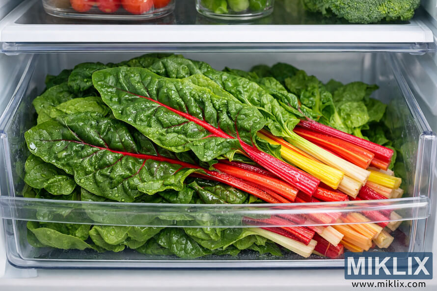 A close-up view of fresh Swiss chard stored neatly in a refrigerator crisper drawer, showing vibrant green leaves and colorful stems.