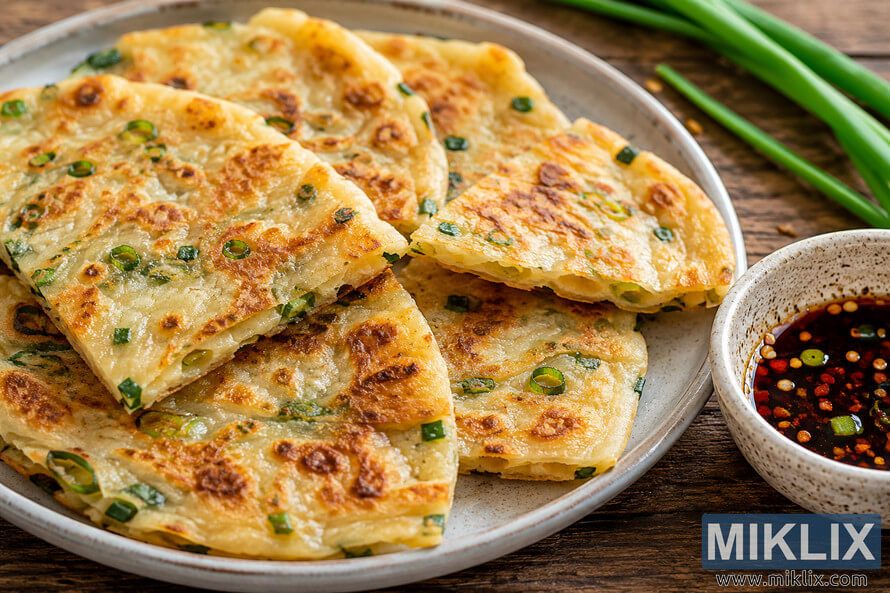 Golden-brown crispy scallion pancakes arranged on a rustic ceramic plate beside a small bowl of dark dipping sauce with chili flakes and sesame seeds on a wooden table.