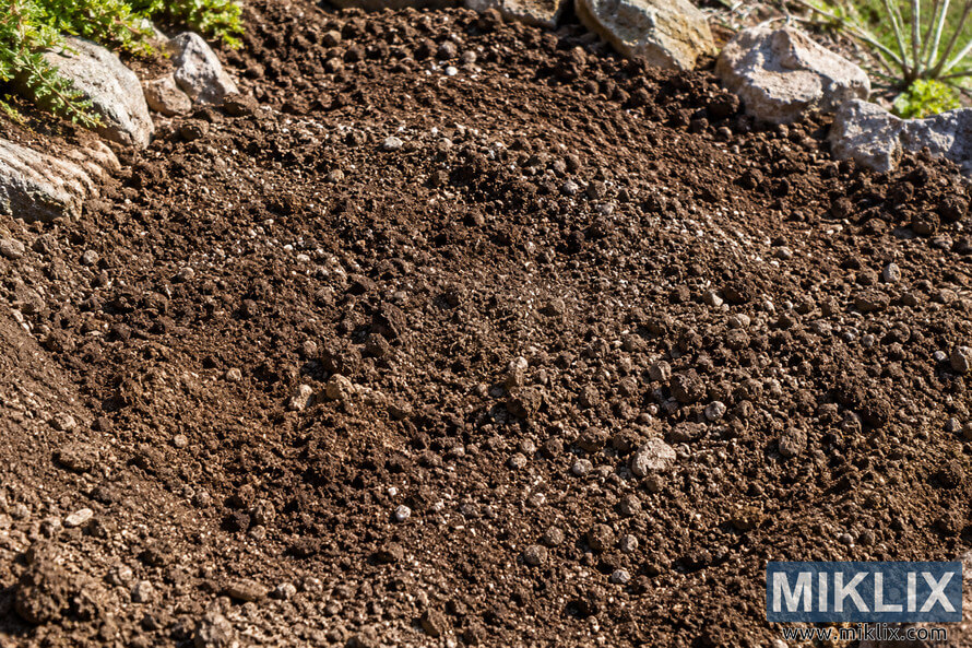 Close-up of loose brown garden soil mixed with small stones and coarse particles, bordered by rocks and sparse green vegetation. Close-up of loose brown garden soil mixed with small stones and coarse particles, bordered by rocks and sparse green vegetation.