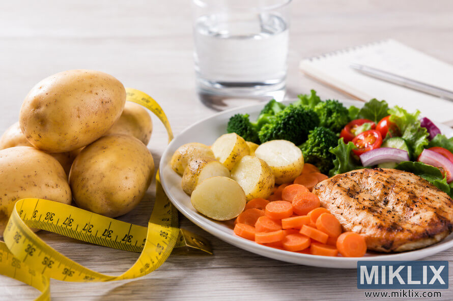 A healthy weight management concept featuring fresh potatoes wrapped with a yellow measuring tape beside a balanced meal of grilled chicken, cooked potatoes, broccoli, carrots, fresh salad, and a glass of water on a light wooden table. A healthy weight management concept featuring fresh potatoes wrapped with a yellow measuring tape beside a balanced meal of grilled chicken, cooked potatoes, broccoli, carrots, fresh salad, and a glass of water on a light wooden table.