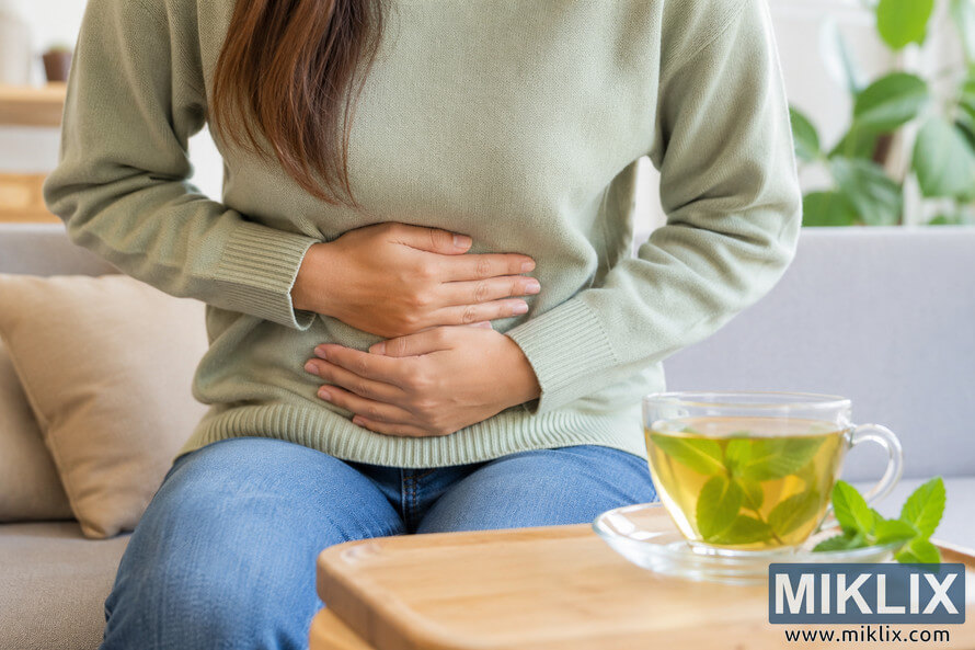 Person sitting on a sofa holding their stomach with both hands while a glass cup of mint herbal tea rests nearby, illustrating digestive wellness and natural comfort in a cozy indoor setting. Person sitting on a sofa holding their stomach with both hands while a glass cup of mint herbal tea rests nearby, illustrating digestive wellness and natural comfort in a cozy indoor setting.