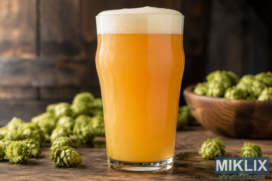 Hazy golden-orange IPA with a creamy foam head in a clear pint glass, surrounded by fresh green hop cones and a wooden bowl of hops on a rustic wooden table.