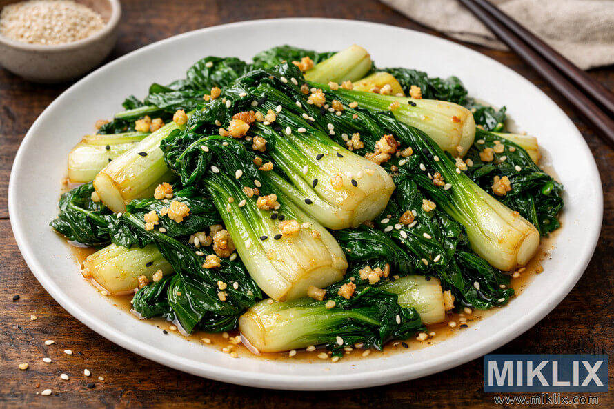 A plated dish of sautéed garlic bok choy garnished with sesame seeds and minced garlic on a white ceramic plate. A plated dish of sautéed garlic bok choy garnished with sesame seeds and minced garlic on a white ceramic plate.