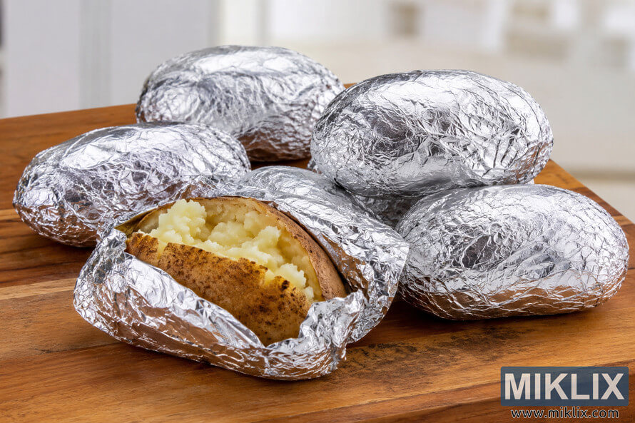 Several baked potatoes wrapped in aluminum foil rest on a wooden cutting board in a softly lit kitchen. One potato is partially unwrapped, revealing its fluffy interior, while the others remain fully wrapped and ready for easy reheating. Several baked potatoes wrapped in aluminum foil rest on a wooden cutting board in a softly lit kitchen. One potato is partially unwrapped, revealing its fluffy interior, while the others remain fully wrapped and ready for easy reheating.