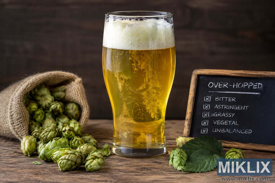 Landscape still life of a golden beer with a foamy head surrounded by abundant green hop cones and leaves, beside a chalkboard labeled âOVER-HOPPEDâ listing bitter, astringent, grassy, vegetal, and unbalanced characteristics.