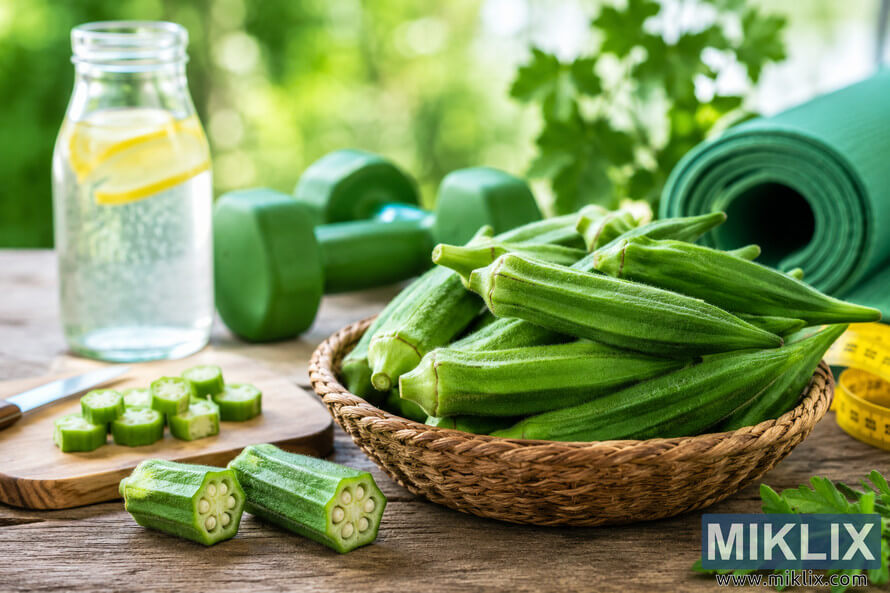 Fresh green okra in a woven basket on a rustic wooden table with sliced okra, sparkling lemon water, a cutting board, green dumbbells, a rolled exercise mat, and a measuring tape against a softly blurred natural background, representing healthy eating and wellness. Fresh green okra in a woven basket on a rustic wooden table with sliced okra, sparkling lemon water, a cutting board, green dumbbells, a rolled exercise mat, and a measuring tape against a softly blurred natural background, representing healthy eating and wellness.
