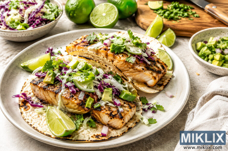 A high-resolution landscape food photograph featuring grilled fish tacos topped with shredded cabbage, purple cabbage, avocado, cilantro, and creamy sauce, served with fresh lime wedges, salsa, and chopped herbs on a light stone countertop.