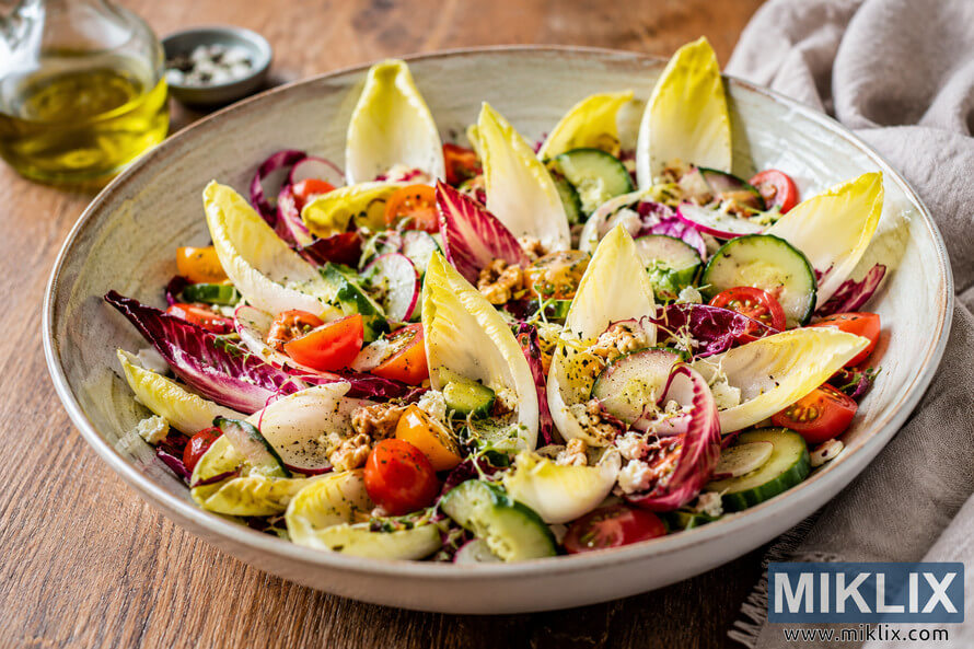 A vibrant healthy salad bowl featuring crisp Belgian endive leaves, cherry tomatoes, cucumber slices, radishes, purple cabbage, mixed greens, crumbled cheese, and walnuts arranged in a rustic ceramic bowl on a wooden table with natural lighting. A vibrant healthy salad bowl featuring crisp Belgian endive leaves, cherry tomatoes, cucumber slices, radishes, purple cabbage, mixed greens, crumbled cheese, and walnuts arranged in a rustic ceramic bowl on a wooden table with natural lighting.