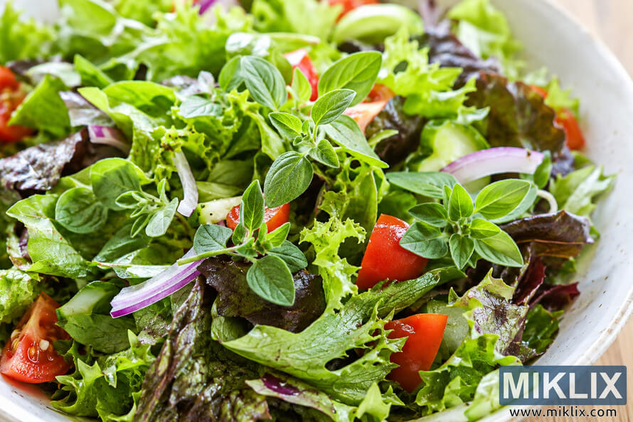 Close-up landscape photograph of a fresh mixed green salad topped with vibrant marjoram sprigs, red onion slices, cucumber, and tomato pieces in a rustic cream-colored bowl.