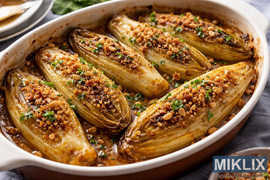 Cooked Belgian endive halves in an oval ceramic serving dish, topped with toasted breadcrumbs, parsley, and pine nuts in a glossy buttery glaze.
