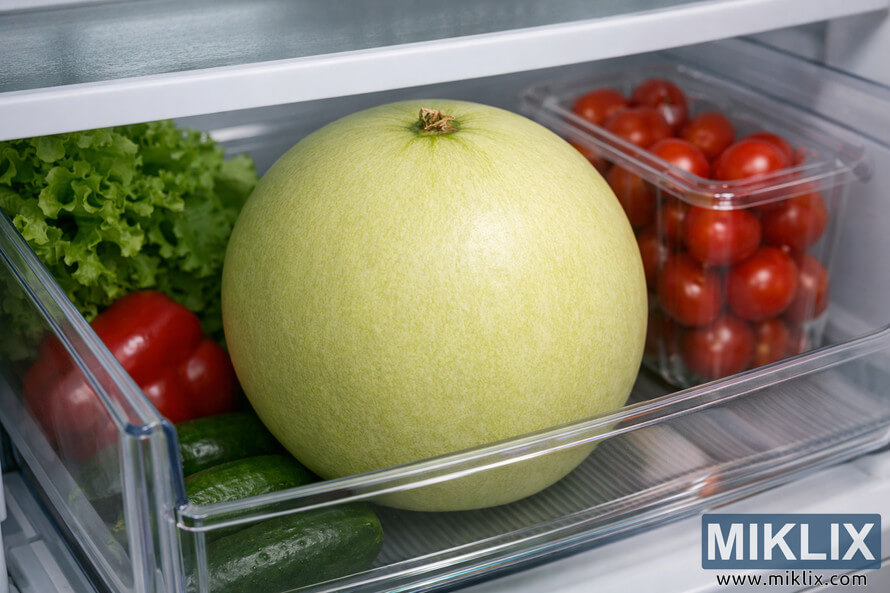 A whole honeydew melon resting in the crisper drawer of a refrigerator surrounded by fresh vegetables. A whole honeydew melon resting in the crisper drawer of a refrigerator surrounded by fresh vegetables.