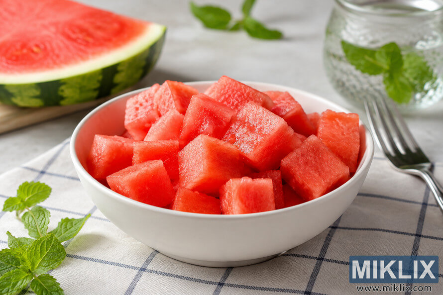 A bright, high-resolution landscape photograph of a white ceramic bowl filled with freshly cut watermelon cubes on a light checkered cloth. Fresh mint sprigs, a sliced watermelon in the softly blurred background, and a glass jar of mint-infused water create a clean, refreshing, and healthy food scene illuminated by natural daylight. A bright, high-resolution landscape photograph of a white ceramic bowl filled with freshly cut watermelon cubes on a light checkered cloth. Fresh mint sprigs, a sliced watermelon in the softly blurred background, and a glass jar of mint-infused water create a clean, refreshing, and healthy food scene illuminated by natural daylight.