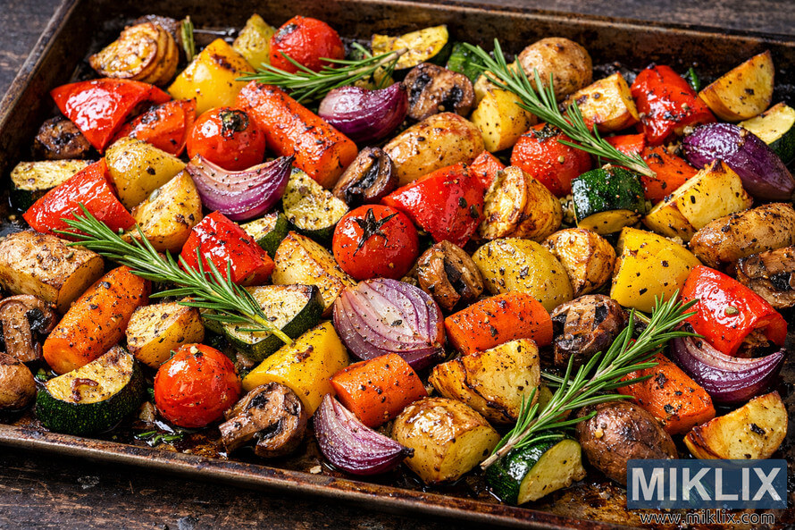 A close-up of colorful roasted vegetables including bell peppers, cherry tomatoes, onions, carrots, zucchini, squash, potatoes, and mushrooms on a dark baking sheet garnished with fresh rosemary sprigs. A close-up of colorful roasted vegetables including bell peppers, cherry tomatoes, onions, carrots, zucchini, squash, potatoes, and mushrooms on a dark baking sheet garnished with fresh rosemary sprigs.