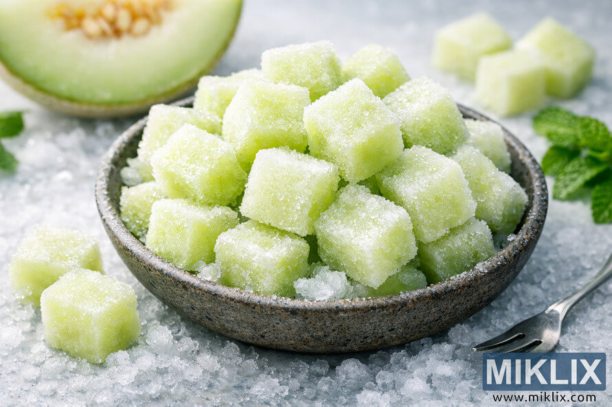 Close-up of frozen honeydew melon cubes piled in a rustic ceramic bowl on a frosty white surface, surrounded by crushed ice, loose melon cubes, fresh mint sprigs, and a silver serving fork, with a blurred honeydew slice and melon chunks in the background. Close-up of frozen honeydew melon cubes piled in a rustic ceramic bowl on a frosty white surface, surrounded by crushed ice, loose melon cubes, fresh mint sprigs, and a silver serving fork, with a blurred honeydew slice and melon chunks in the background.