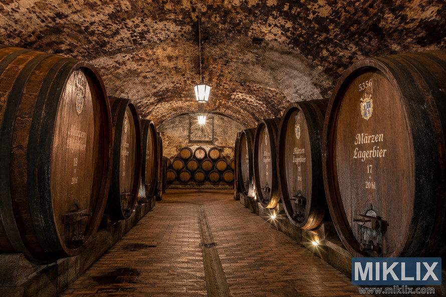 Historic German lagering cellar with rows of wooden Märzen lager barrels beneath vaulted stone ceilings illuminated by warm lantern light. Historic German lagering cellar with rows of wooden Märzen lager barrels beneath vaulted stone ceilings illuminated by warm lantern light.