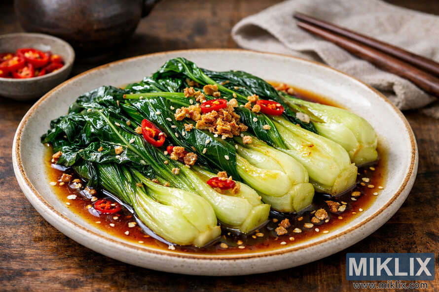 A close-up three-quarter view of a beautifully plated sautéed bok choy dish on an off-white ceramic plate with a rustic brown rim, drizzled with glossy savory sauce and garnished with crispy garlic bits, thin red chili slices, and white sesame seeds, set on a wooden table with small dishes of chili and sesame seeds and a pair of wooden chopsticks resting on a beige linen cloth in the blurred background. A close-up three-quarter view of a beautifully plated sautéed bok choy dish on an off-white ceramic plate with a rustic brown rim, drizzled with glossy savory sauce and garnished with crispy garlic bits, thin red chili slices, and white sesame seeds, set on a wooden table with small dishes of chili and sesame seeds and a pair of wooden chopsticks resting on a beige linen cloth in the blurred background.