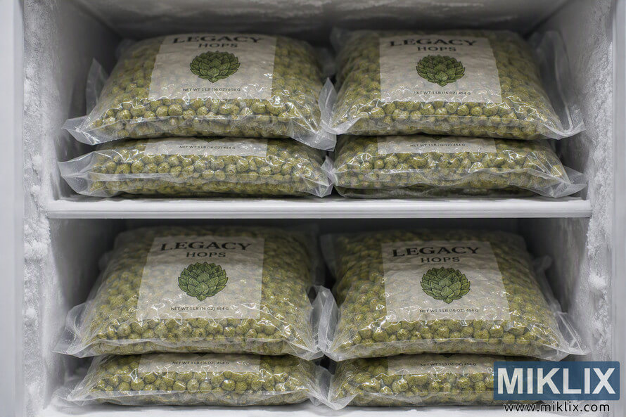 Multiple transparent vacuum-sealed bags labeled Legacy Hops are neatly stacked on two freezer shelves inside a frost-covered chest freezer. The green hop pellets are visible through the packaging, and the frozen storage environment highlights optimal preservation conditions for brewing ingredients. Multiple transparent vacuum-sealed bags labeled Legacy Hops are neatly stacked on two freezer shelves inside a frost-covered chest freezer. The green hop pellets are visible through the packaging, and the frozen storage environment highlights optimal preservation conditions for brewing ingredients.
