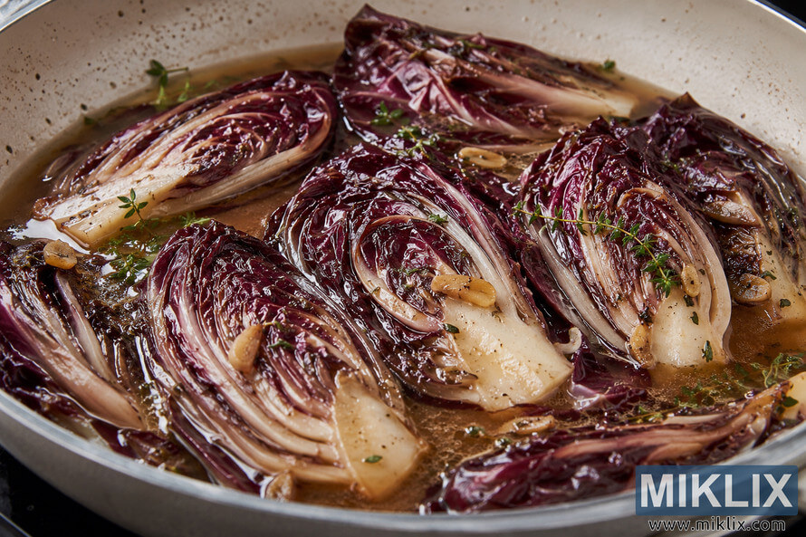 A close-up landscape photograph of several wedges of purple radicchio gently braising in a shallow herb-infused broth with sliced garlic and fresh herbs inside a light-colored skillet. The tender leaves glisten under soft natural light, highlighting their rich burgundy hues, creamy white ribs, and delicate textures. A close-up landscape photograph of several wedges of purple radicchio gently braising in a shallow herb-infused broth with sliced garlic and fresh herbs inside a light-colored skillet. The tender leaves glisten under soft natural light, highlighting their rich burgundy hues, creamy white ribs, and delicate textures.
