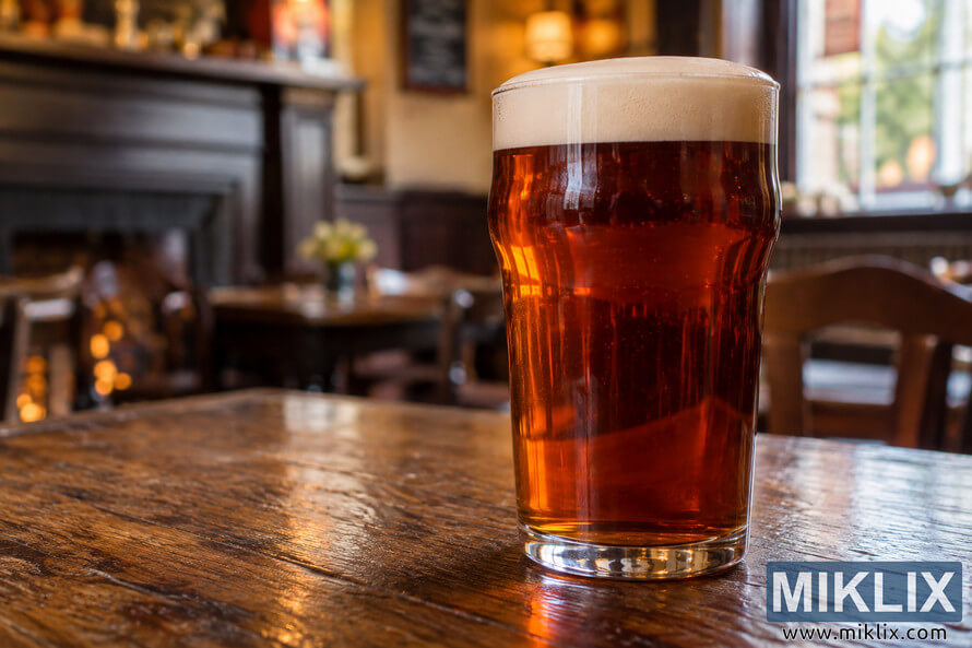 Freshly poured unbranded pint of copper-colored British bitter with a thick, creamy, well-retained head on a rustic wooden table inside a warmly lit traditional pub.