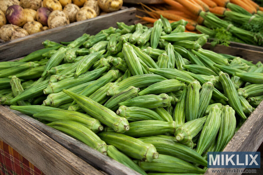 Close-up of freshly harvested green okra piled in a rustic wooden crate at a farmers market, with potatoes, carrots, and leafy vegetables softly blurred in the background under natural daylight. Close-up of freshly harvested green okra piled in a rustic wooden crate at a farmers market, with potatoes, carrots, and leafy vegetables softly blurred in the background under natural daylight.