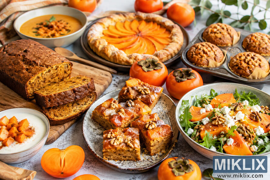 A warm autumn-inspired food photography scene featuring baked persimmon desserts, muffins, loaf cake, tart, salad, and creamy soup arranged on a rustic table with fresh ripe persimmons and soft natural lighting.