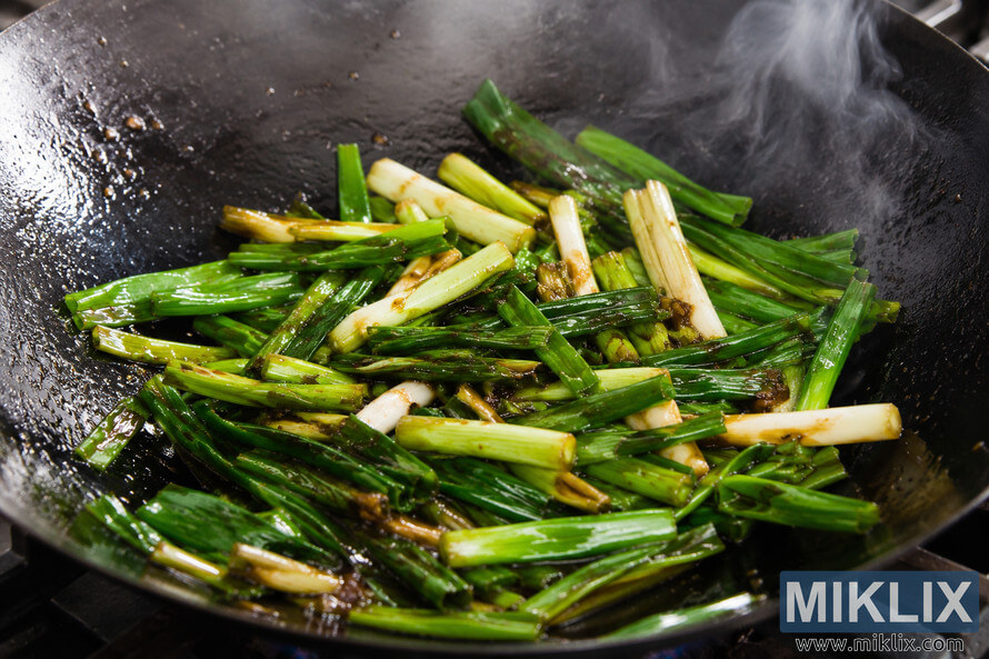 Close-up landscape food photograph of fresh green scallions being stir-fried in a dark seasoned wok with glossy soy sauce. Steam rises from the hot pan while the scallions glisten with oil and caramelized sauce under dramatic kitchen lighting.