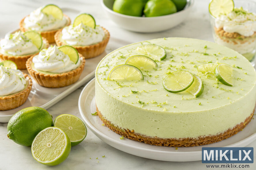 Landscape food photograph featuring a creamy lime cheesecake topped with fresh lime slices, mini lime tartlets with whipped topping, a layered lime parfait, whole and halved fresh limes, and a bowl of limes on a bright marble countertop.