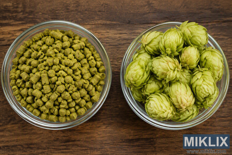 Top-down comparison image showing two clear glass bowls on a rustic wooden surface. The bowl on the left contains green hop pellets, while the bowl on the right contains fresh whole cone hops. The image highlights the visual differences in shape, texture, and appearance between processed hop pellets and unprocessed whole hop cones.