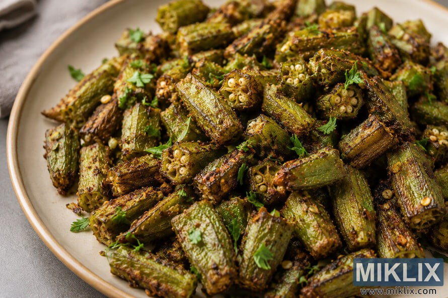 A close-up landscape photograph of roasted okra seasoned with herbs, black pepper, chili flakes, and spices, served on a rustic beige ceramic plate with a softly blurred neutral background. A close-up landscape photograph of roasted okra seasoned with herbs, black pepper, chili flakes, and spices, served on a rustic beige ceramic plate with a softly blurred neutral background.