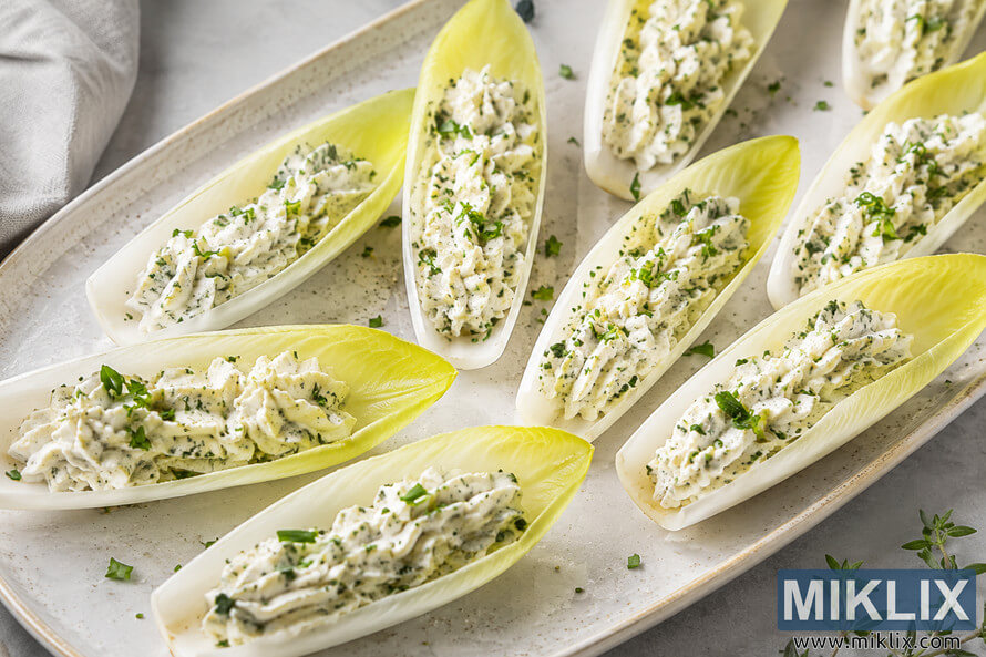 Close-up overhead view of fresh Belgian endive leaves arranged on a white ceramic serving platter, each filled with a creamy herbed cheese spread and garnished with chopped green herbs on a light neutral background. Close-up overhead view of fresh Belgian endive leaves arranged on a white ceramic serving platter, each filled with a creamy herbed cheese spread and garnished with chopped green herbs on a light neutral background.