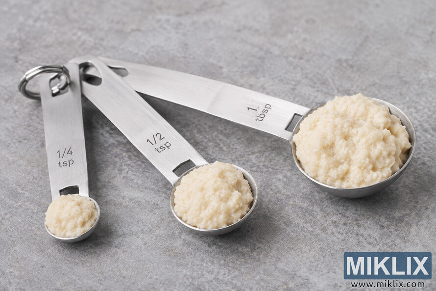 Close-up landscape photo of stainless steel measuring spoons containing prepared horseradish portions arranged diagonally on a textured gray stone background.