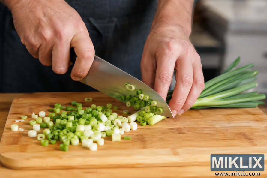 Close-up landscape photo of fresh scallions being chopped with a chef’s knife on a wooden cutting board, showing proper slicing technique in a warm kitchen setting.