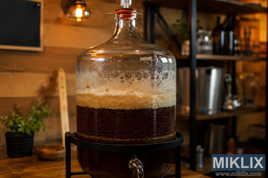 A warmly lit homebrewing workspace featuring a large glass fermentation vessel filled with dark strong ale during active fermentation. A thick foamy krausen crowns the liquid, condensation coats the glass, and an airlock sits at the top. Wooden walls, brewing equipment, shelves, and soft amber lighting create a cozy, handcrafted atmosphere.