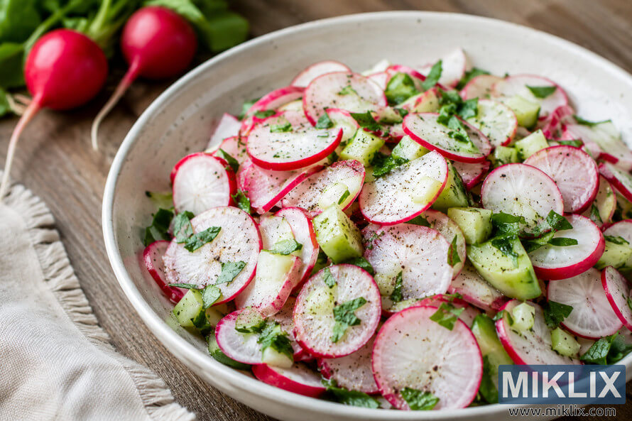 High-resolution landscape photograph of a healthy radish salad served in a rustic ceramic bowl with sliced radishes, cucumber, fresh herbs, cracked black pepper, whole radishes in the softly blurred background, and natural daylight on a wooden table.