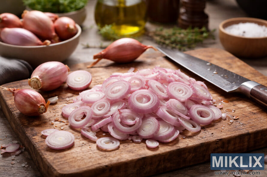 Thinly sliced shallots arranged on a wooden cutting board with a chefâs knife and whole shallots in a warm kitchen setting.