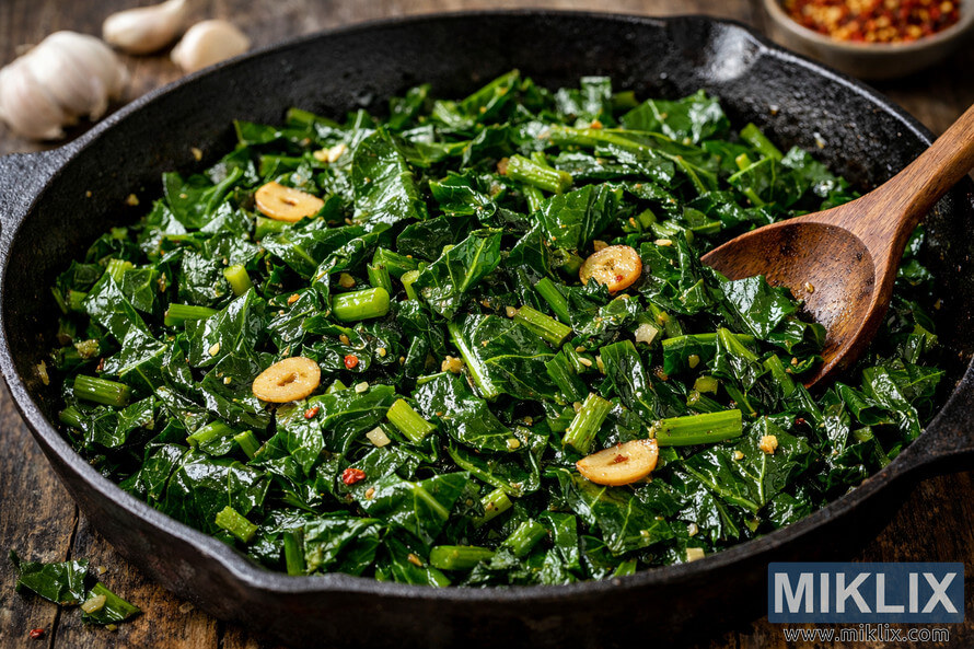 A close-up view of sautéed collard greens with golden garlic slices in a black cast iron skillet, glistening with oil and red pepper flakes, with a wooden spoon resting on the side.