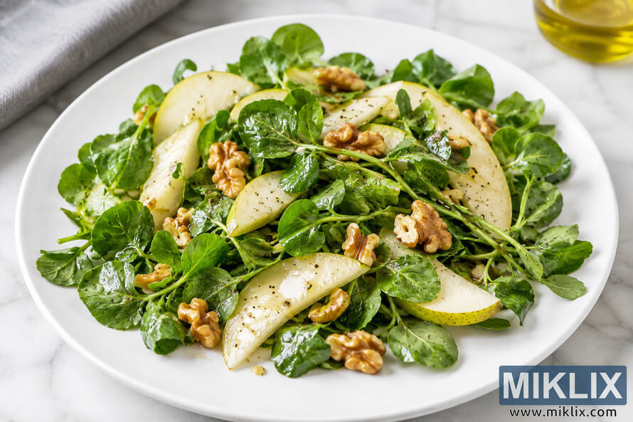A fresh watercress and pear salad served on a white plate, topped with walnut pieces and lightly dressed, photographed in soft natural light on a marble surface. A fresh watercress and pear salad served on a white plate, topped with walnut pieces and lightly dressed, photographed in soft natural light on a marble surface.
