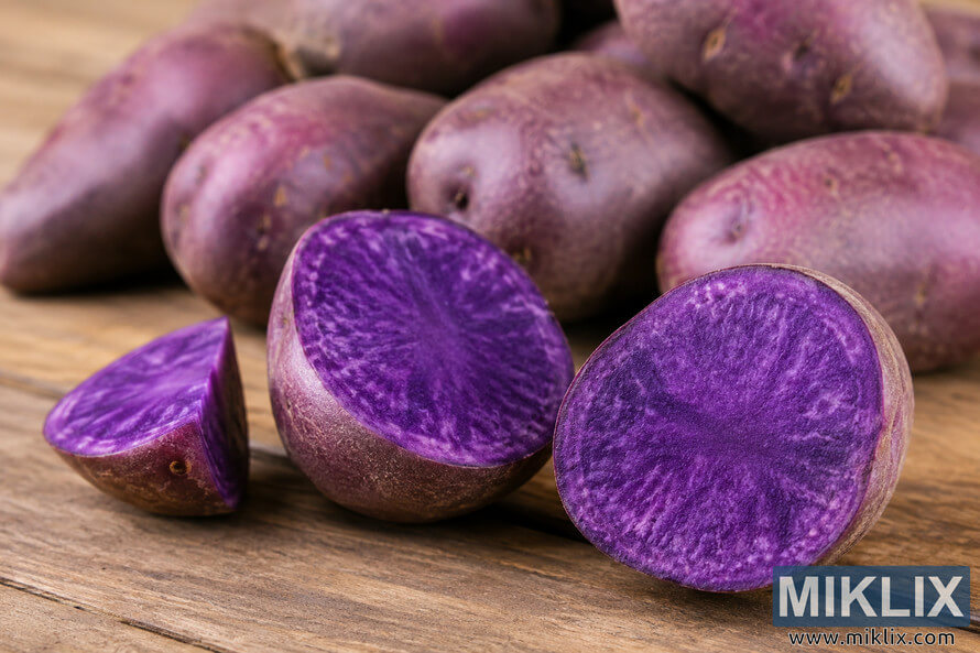 Close-up landscape photograph of whole and halved purple potatoes displayed on a rustic wooden table, highlighting their vibrant violet flesh rich in anthocyanins with a softly blurred background. Close-up landscape photograph of whole and halved purple potatoes displayed on a rustic wooden table, highlighting their vibrant violet flesh rich in anthocyanins with a softly blurred background.