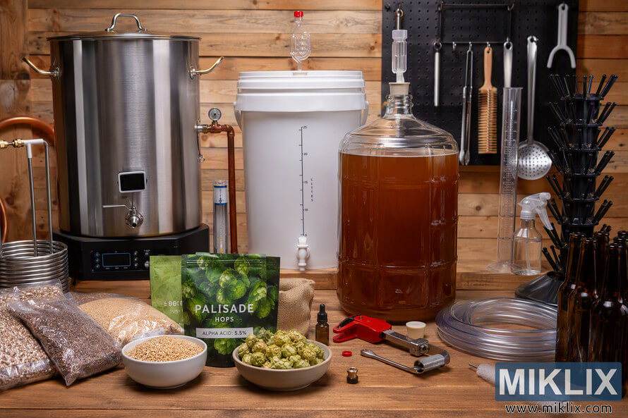 Comprehensive homebrewing workstation displayed on a wooden workbench, featuring a stainless-steel brewing kettle, fermentation bucket, glass carboy filled with amber liquid, grain ingredients, hop pellets with Palisade hops packaging, tubing, bottles, bottle-drying rack, measuring tools, cleaning supplies, and brewing accessories arranged in a rustic workshop setting. Comprehensive homebrewing workstation displayed on a wooden workbench, featuring a stainless-steel brewing kettle, fermentation bucket, glass carboy filled with amber liquid, grain ingredients, hop pellets with Palisade hops packaging, tubing, bottles, bottle-drying rack, measuring tools, cleaning supplies, and brewing accessories arranged in a rustic workshop setting.