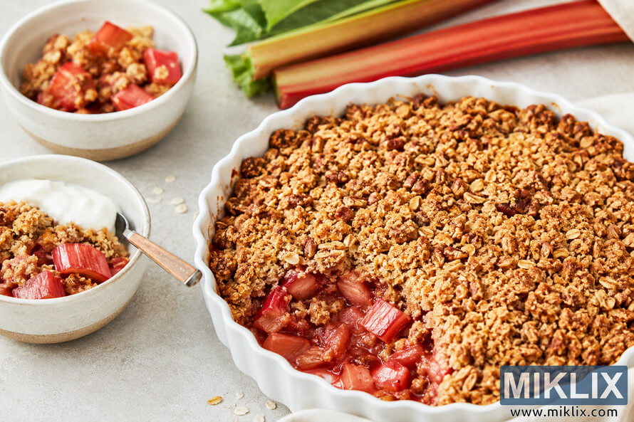 A bright rustic food scene featuring healthy rhubarb desserts without added sugar, including a baked rhubarb oat crumble in a white ceramic dish surrounded by small bowls of fruit crumble and yogurt on a light countertop with fresh rhubarb stalks in the background. A bright rustic food scene featuring healthy rhubarb desserts without added sugar, including a baked rhubarb oat crumble in a white ceramic dish surrounded by small bowls of fruit crumble and yogurt on a light countertop with fresh rhubarb stalks in the background.