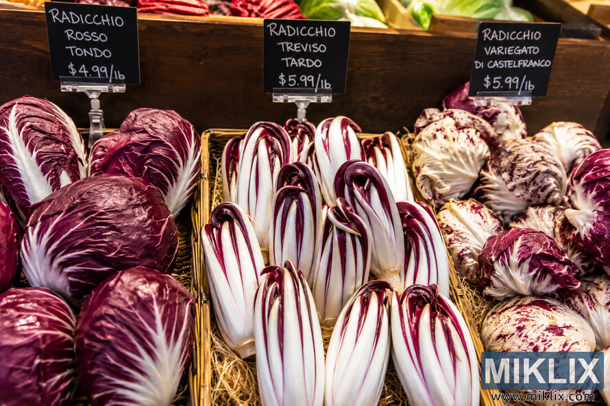 A warmly lit specialty grocery store produce display featuring multiple varieties of radicchio arranged in rustic baskets and wooden crates with handwritten chalkboard signs, creating an inviting market atmosphere without any visible brands or people. A warmly lit specialty grocery store produce display featuring multiple varieties of radicchio arranged in rustic baskets and wooden crates with handwritten chalkboard signs, creating an inviting market atmosphere without any visible brands or people.