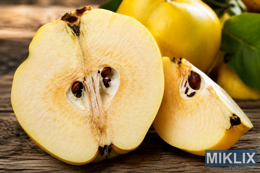 Close-up landscape photograph of a freshly sliced quince on a rustic wooden surface, revealing the pale golden flesh, seed chambers, fibrous core, and natural texture of the fruit with whole quinces and green leaves softly blurred in the background. Close-up landscape photograph of a freshly sliced quince on a rustic wooden surface, revealing the pale golden flesh, seed chambers, fibrous core, and natural texture of the fruit with whole quinces and green leaves softly blurred in the background.
