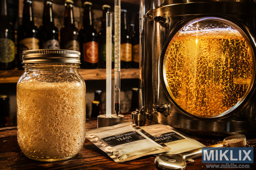 Close-up of a warmly lit brewery showing an active fermentation vessel with bubbling golden liquid, a mason jar containing a lively yeast starter, a hydrometer, generic yeast packets, brewing tools, and a rustic wooden shelf with unbranded beer bottles in the softly blurred background.