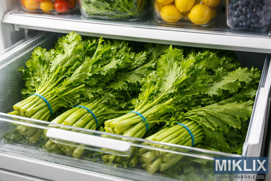 Bundles of fresh mustard greens stored in a refrigerator crisper drawer, with tomatoes, lemons, and blueberries visible on the shelf above.